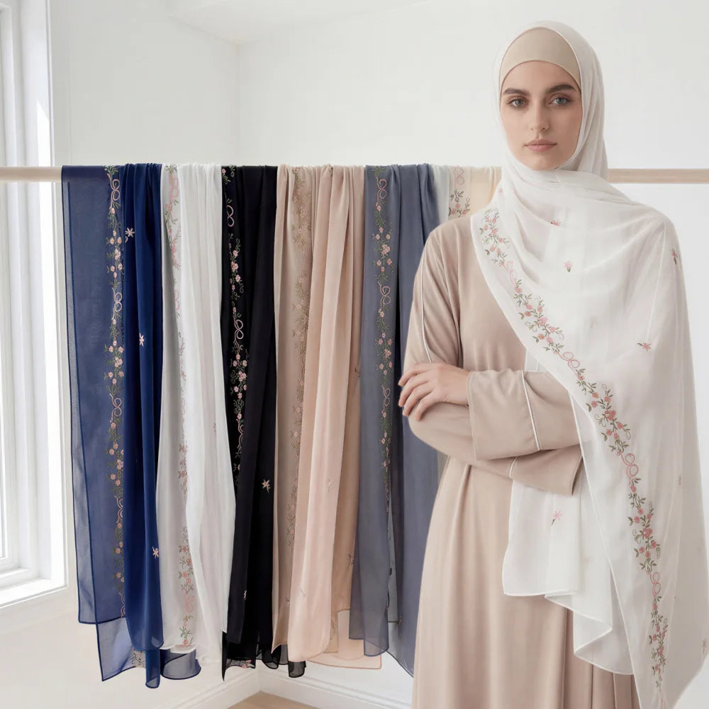 A woman wearing a beige dress and a white hijab with floral embroidery, standing in front of a rack of chiffon shawls.