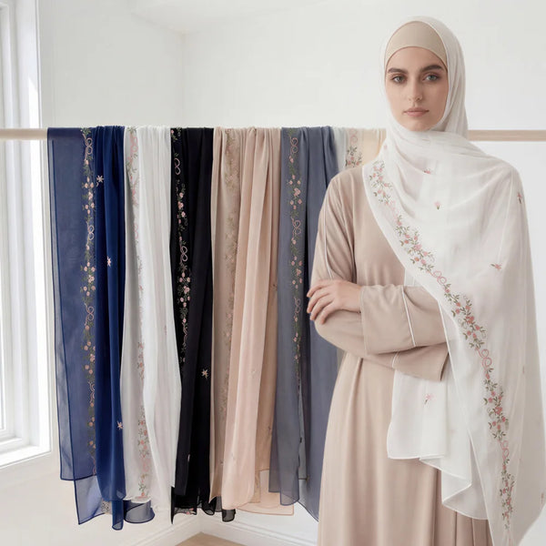 A woman wearing a beige dress and a white hijab with floral embroidery, standing in front of a rack of chiffon shawls.