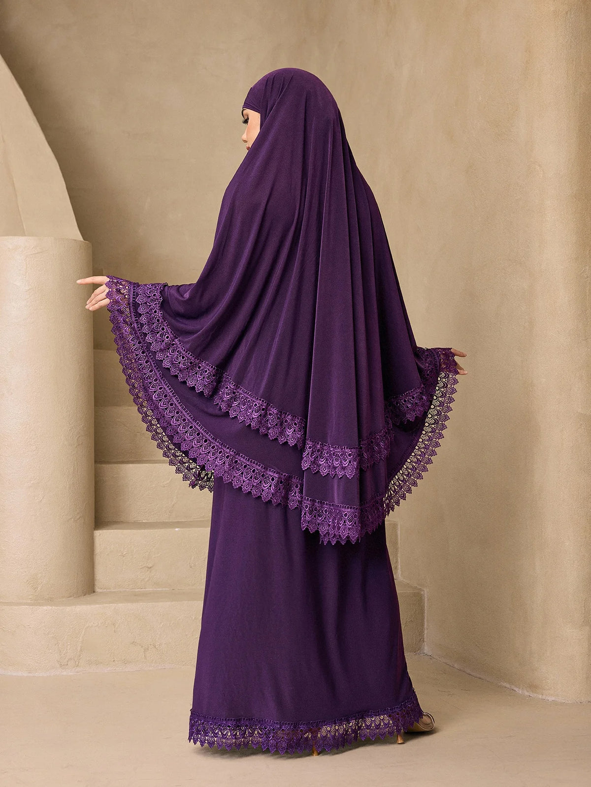 Eine Frau in einem lila, mehrlagigen Spitzen-Khimar-Abaya-Set steht neben Steinstufen.