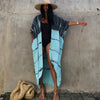 Eine Frau mit blau-gemustertem Kimono-Überwurf über einem schwarzen Kleid und Strohhut steht vor einer grauen Wand.