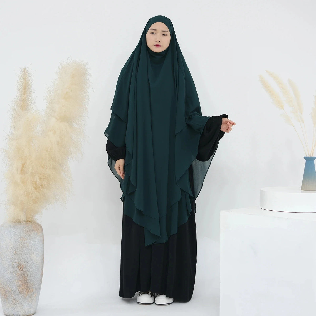 Eine Frau mit einem dunkelgrünen, zweilagigen Chiffon-Niqab und schwarzer Kleidung vor weißem Hintergrund.