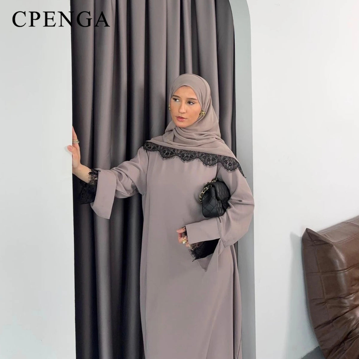 Una mujer vistiendo un modesto traje de abaya gris con detalles de encaje negro, de pie frente a una cortina oscura.