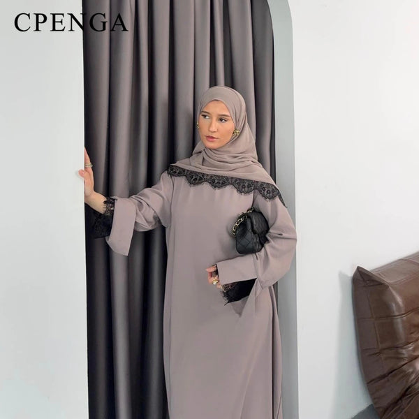 A woman wearing a modest gray abaya set with black lace detailing, standing in front of a dark curtain.