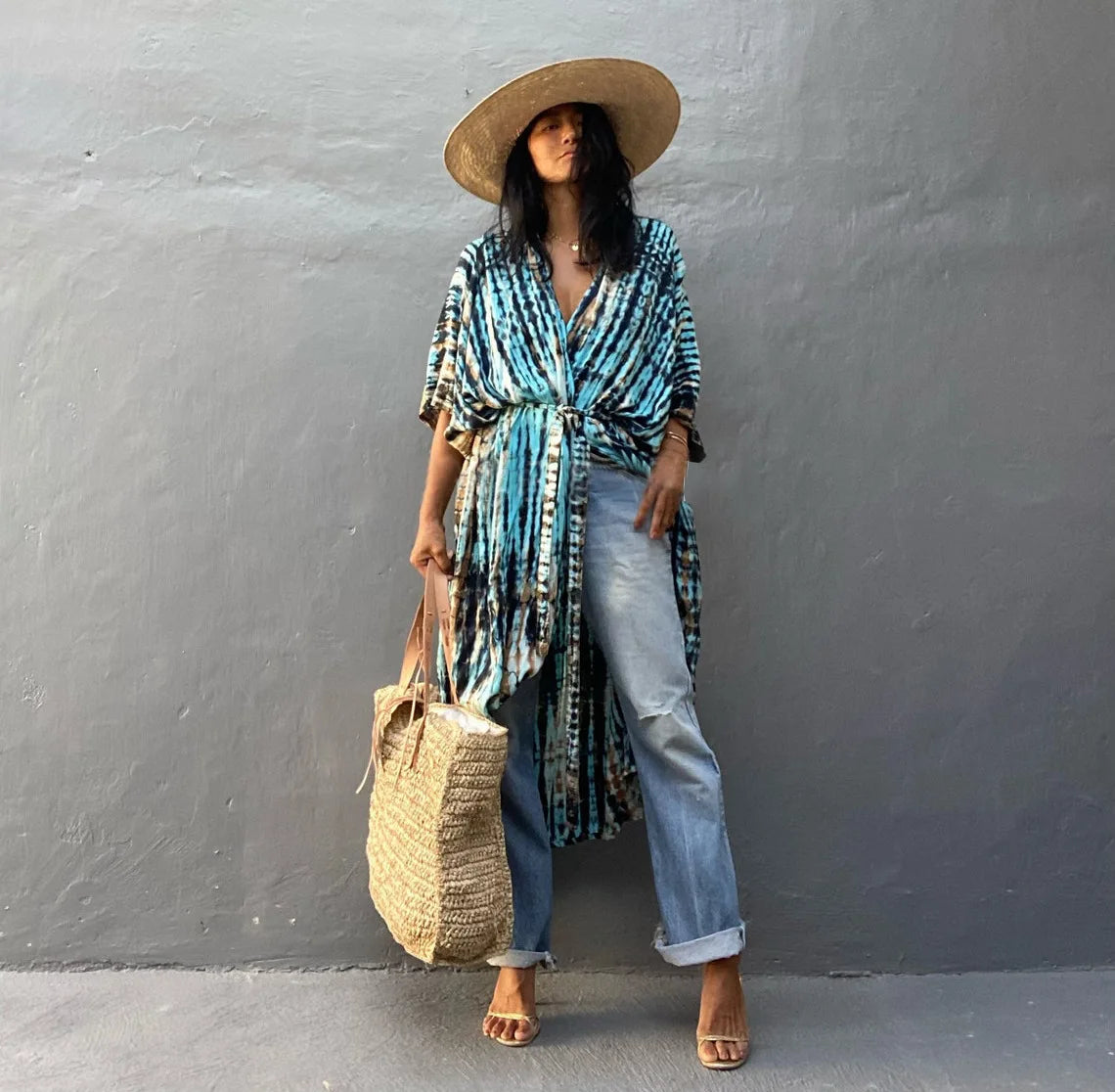 Eine Frau in blau-weiß gemustertem Kimono-Überkleid mit hellgewaschenen Jeans und einer Strohtasche, vor einem Stein