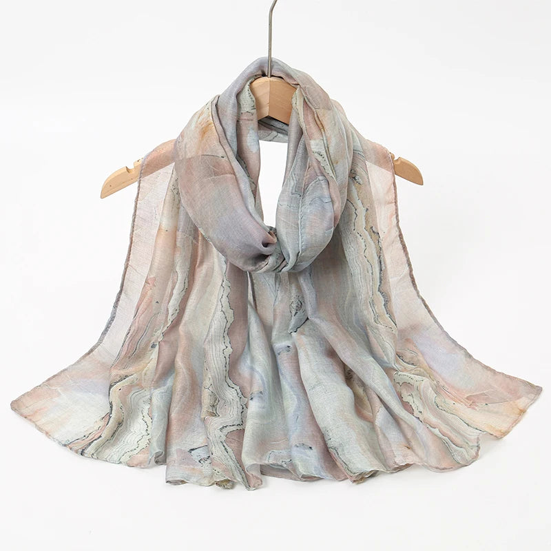 Una bufanda transparente y fluida con estampados de acuarela en rosas suaves y grises, colgada en una percha de madera.