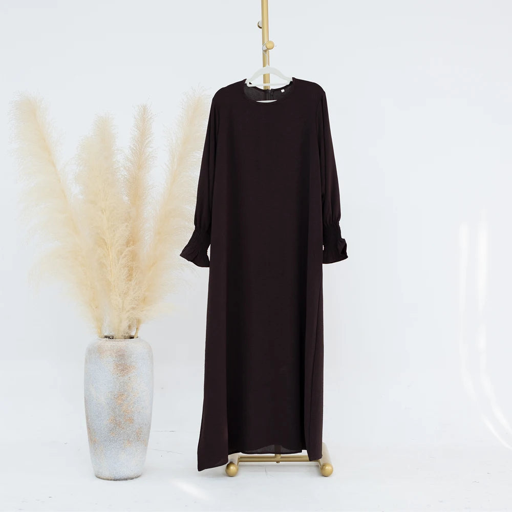 Vestido abaya largo y oscuro marrón, de corte holgado, colgado en percha contra fondo blanco.