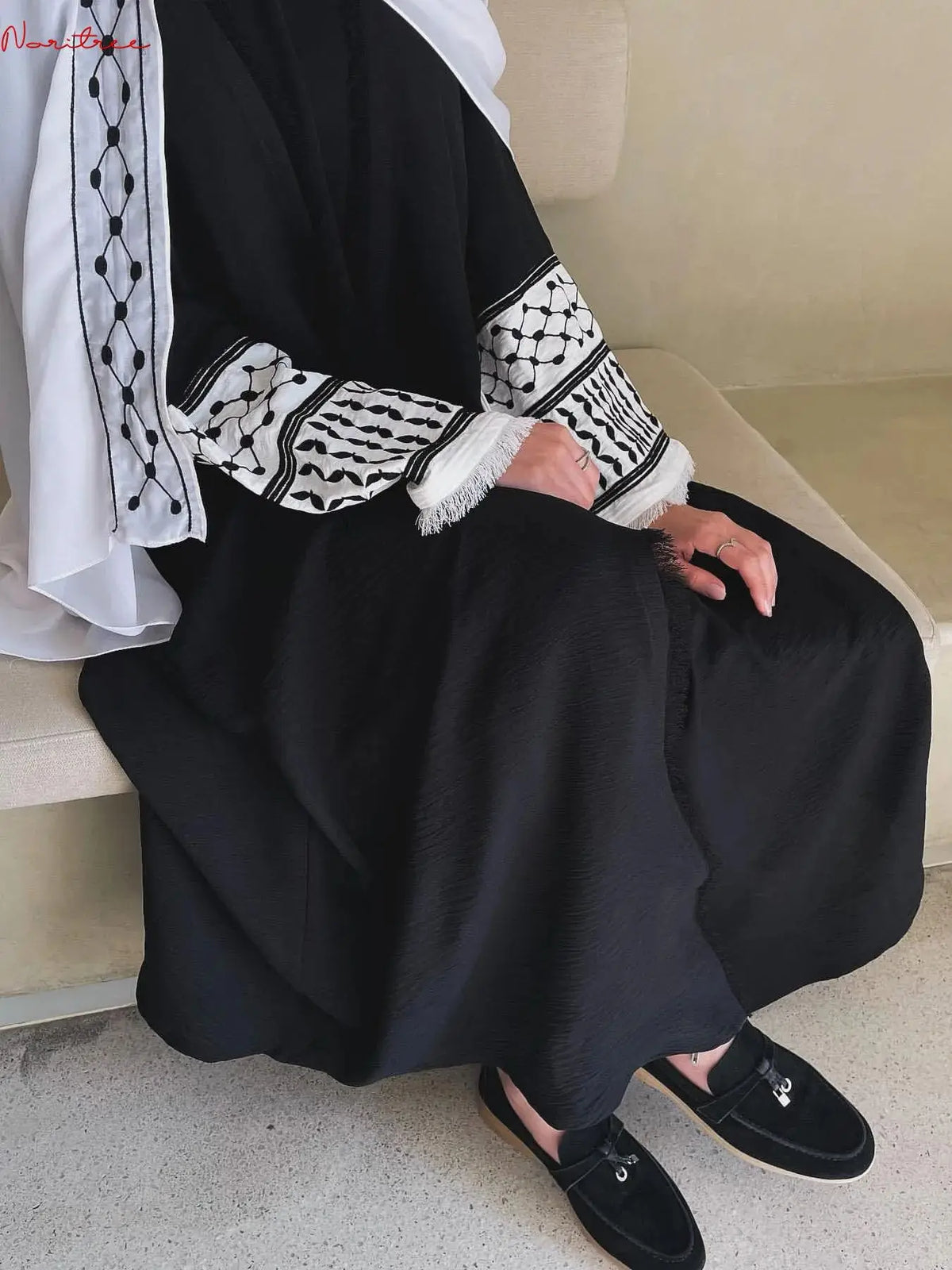 Une femme portant une élégante abaya noire à broderies blanches et un hijab blanc, assise à l'intérieur.