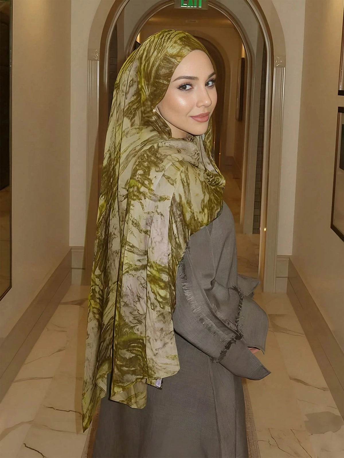 A woman wearing an elegant olive green watercolor print modal scarf and a matching coat in a hallway.