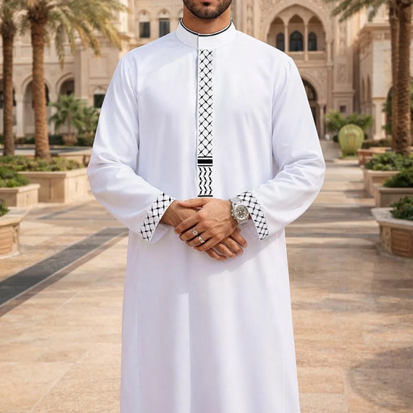 A man wearing a white embroidered jubba thobe kaftan with black and white detailing in an outdoor courtyard setting.
