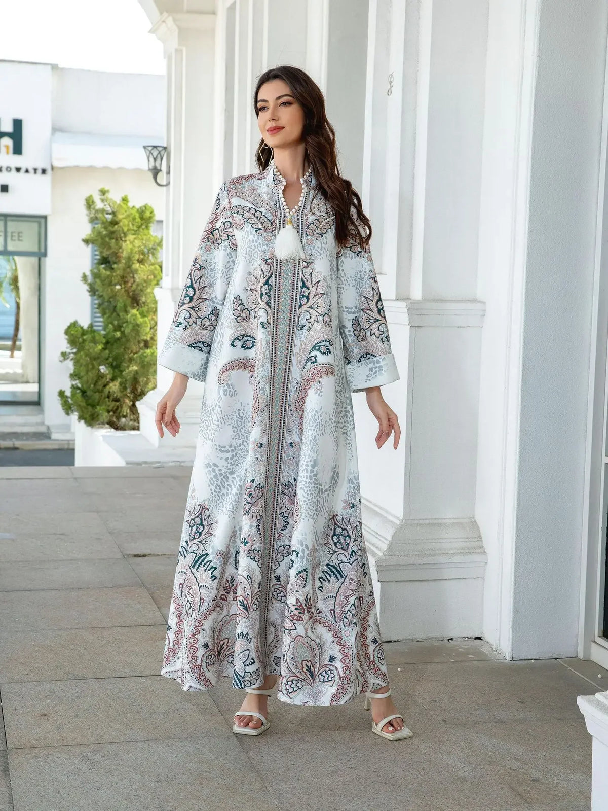 Une femme portant une élégante abaya longue perlée de diamants à motif floral blanc et bleu, debout à l'extérieur.