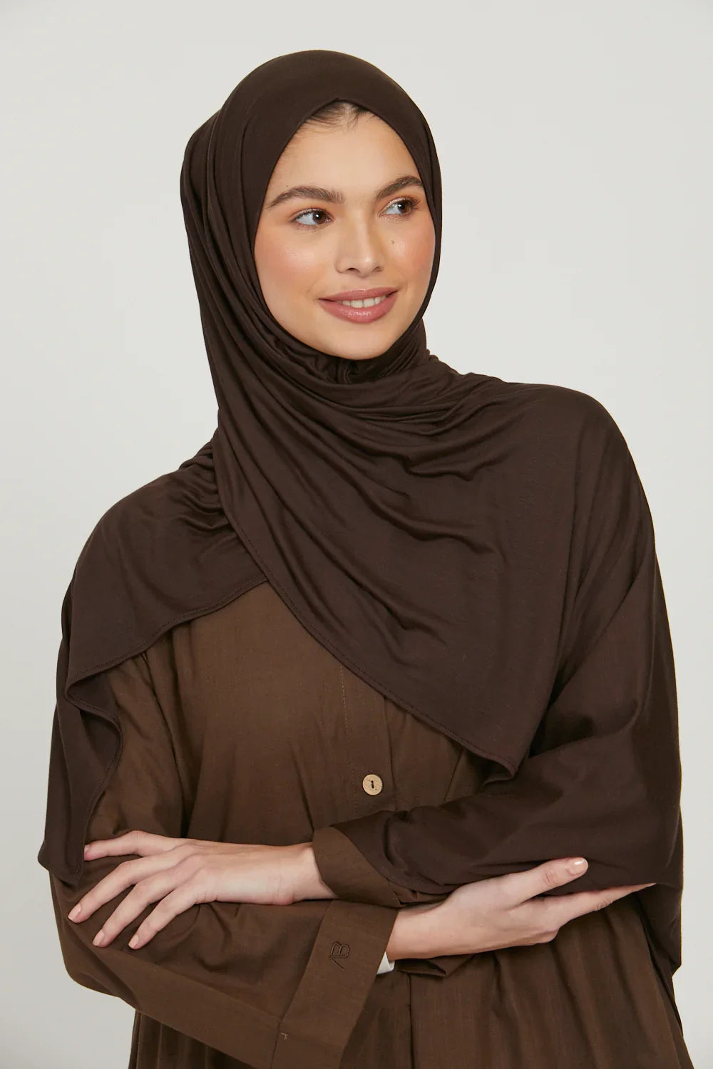 Eine Frau trägt einen einfarbigen braunen Jersey-Hijab und ein passendes Oberteil, blickt zur Seite.