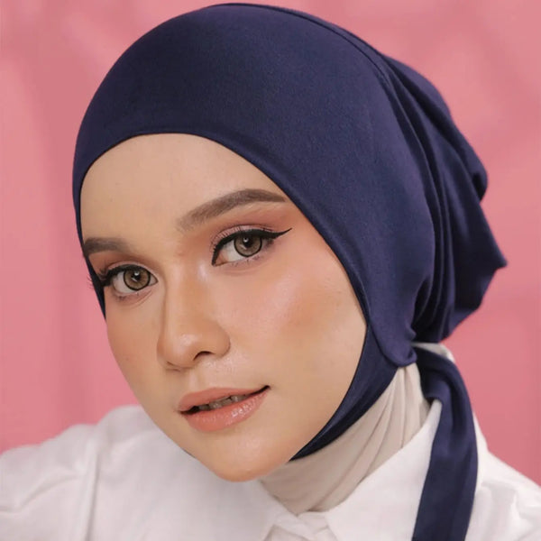 A woman wearing a navy blue inner hijab cap scarf and white top, looking directly at the camera.