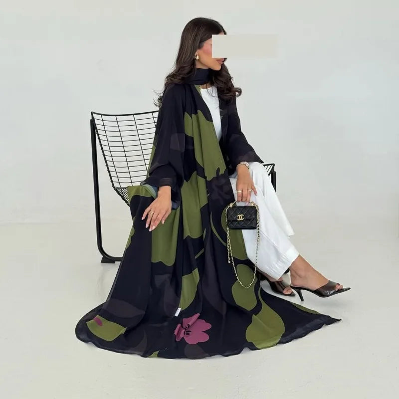 Una mujer modela un conjunto de abaya de gasa negra y verde oliva con pantalones blancos y un bolso negro.