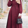 Una mujer vistiendo un vestido largo holgado color granate y hijab beige sostiene un bolso negro al aire libre.