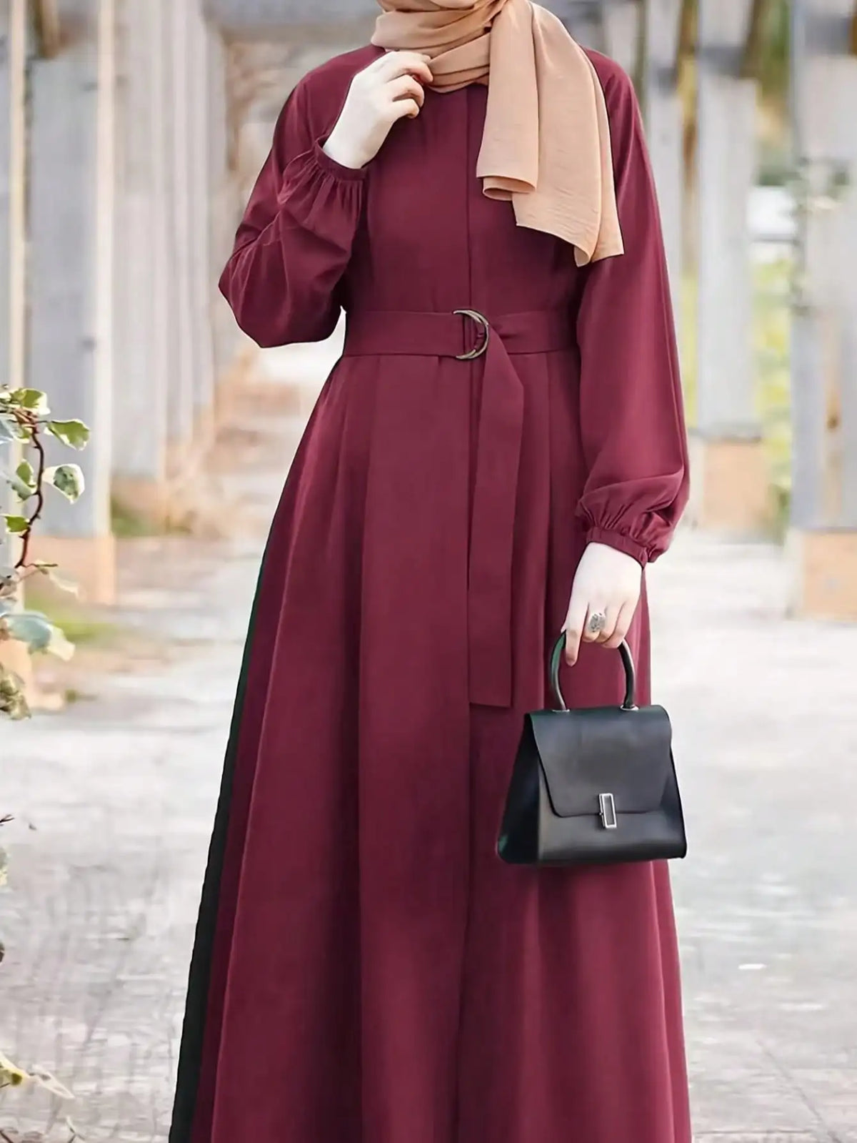 Una mujer vistiendo un vestido largo holgado color granate y hijab beige sostiene un bolso negro al aire libre.
