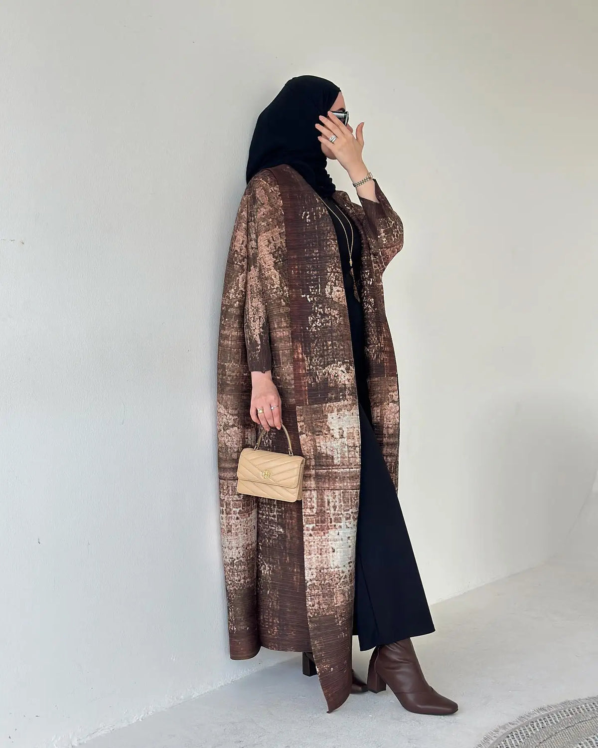 Une femme portant une abaya marron à motifs et un hijab noir, tenant un sac à main beige clair.