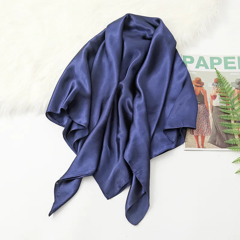 A draped navy blue silk satin hijab scarf lying on a white surface next to a fluffy white rug and a magazine.