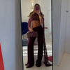 A woman wearing a deep purple sheer mesh poncho cape and matching trousers, holding a clutch, is photographed in a full-lengt