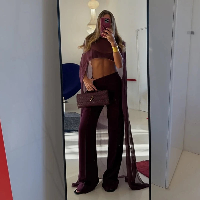 A woman wearing a deep purple sheer mesh poncho cape and matching trousers, holding a clutch, is photographed in a full-lengt