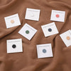 Una colección de cinco pines magnéticos redondos para hijab en varios colores sobre tela marrón.