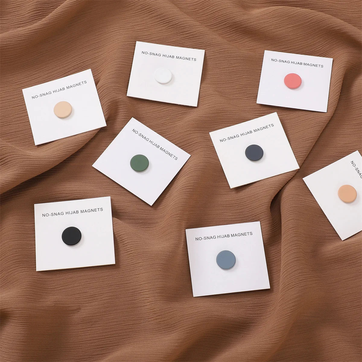 Una colección de cinco pines magnéticos redondos para hijab en varios colores sobre tela marrón.