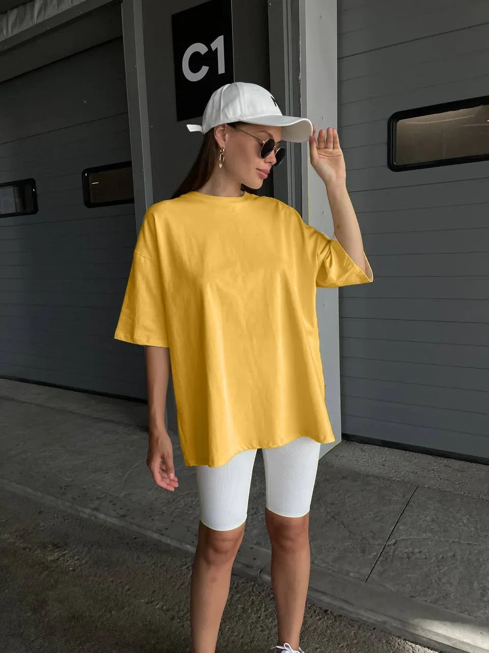 Una mujer con una camiseta básica amarilla grande de algodón y pantalones blancos está frente a una puerta gris.