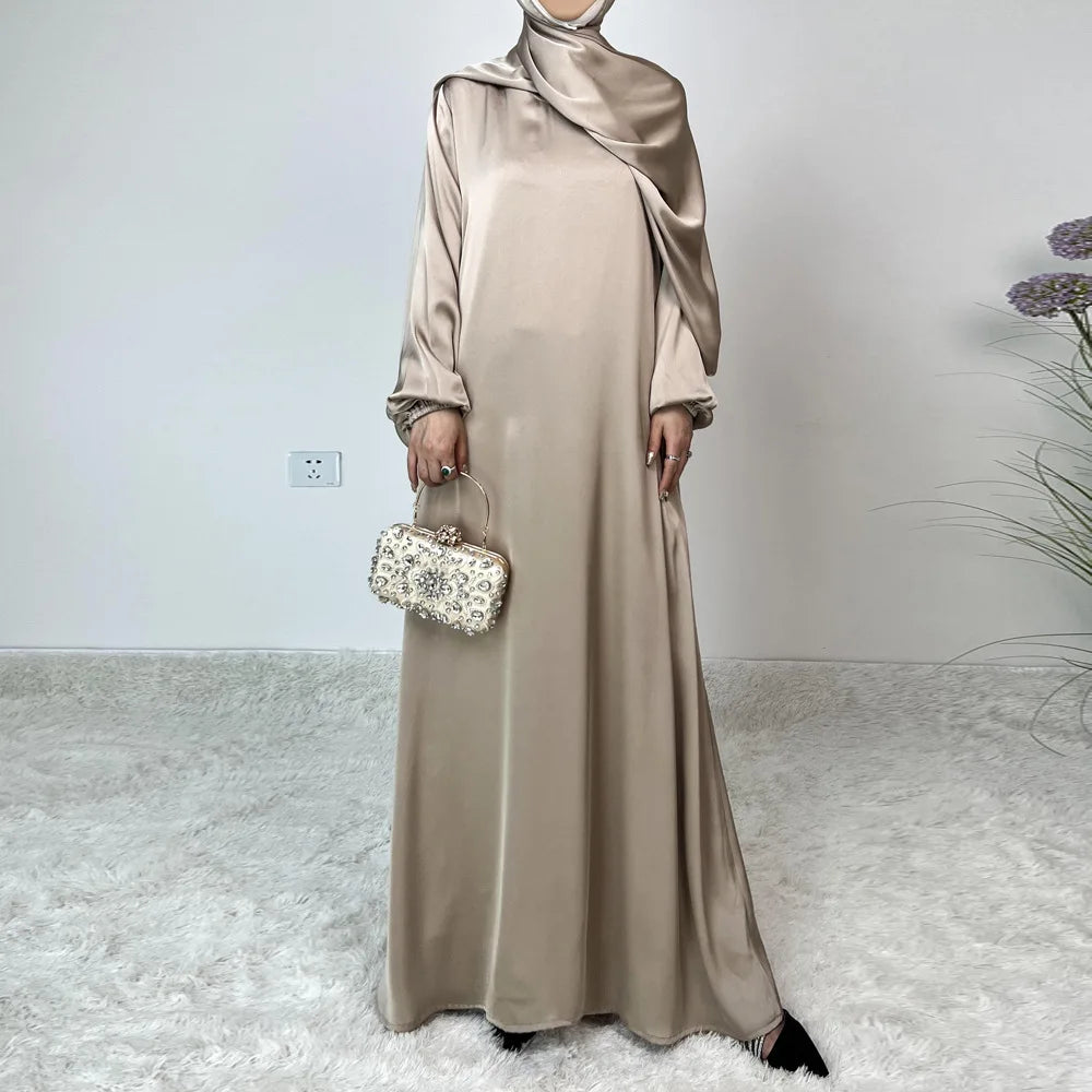 Modelo vistiendo un abaya de satén beige hasta el suelo con hijab a juego y sosteniendo un clutch pequeño.