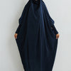 Eine Frau in einem dunkelblauen, bescheidenen Abaya mit gerafften Ärmeln und passender Hijab vor einer weißen Wand.