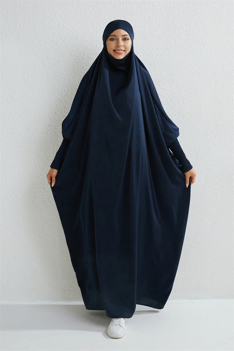 Eine Frau in einem dunkelblauen, bescheidenen Abaya mit gerafften Ärmeln und passender Hijab vor einer weißen Wand.