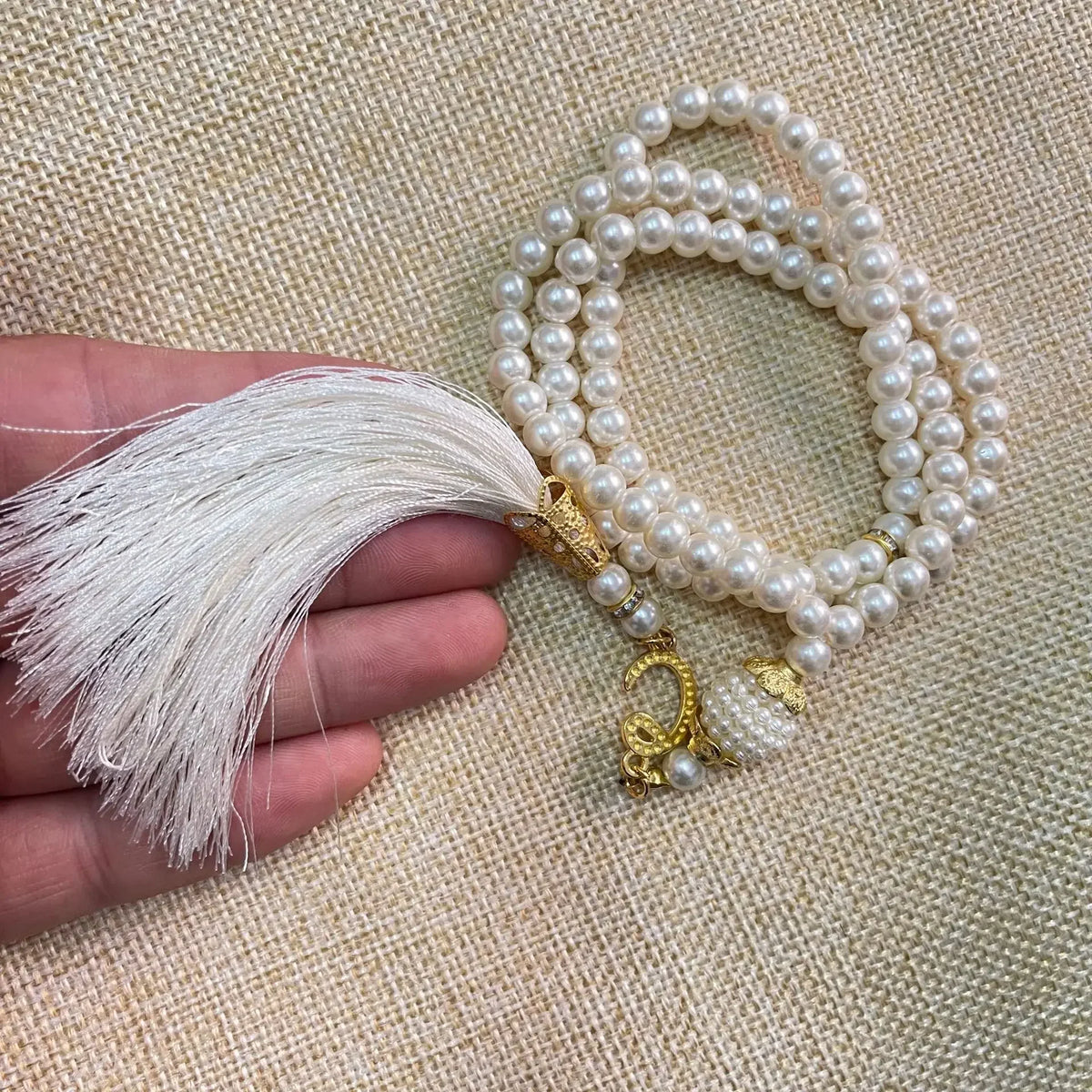 Eine Hand hält ein weißes Perlenrosary-Armband mit Fransen und goldener Anhänger vor einem beigeen Stoffhintergrund.