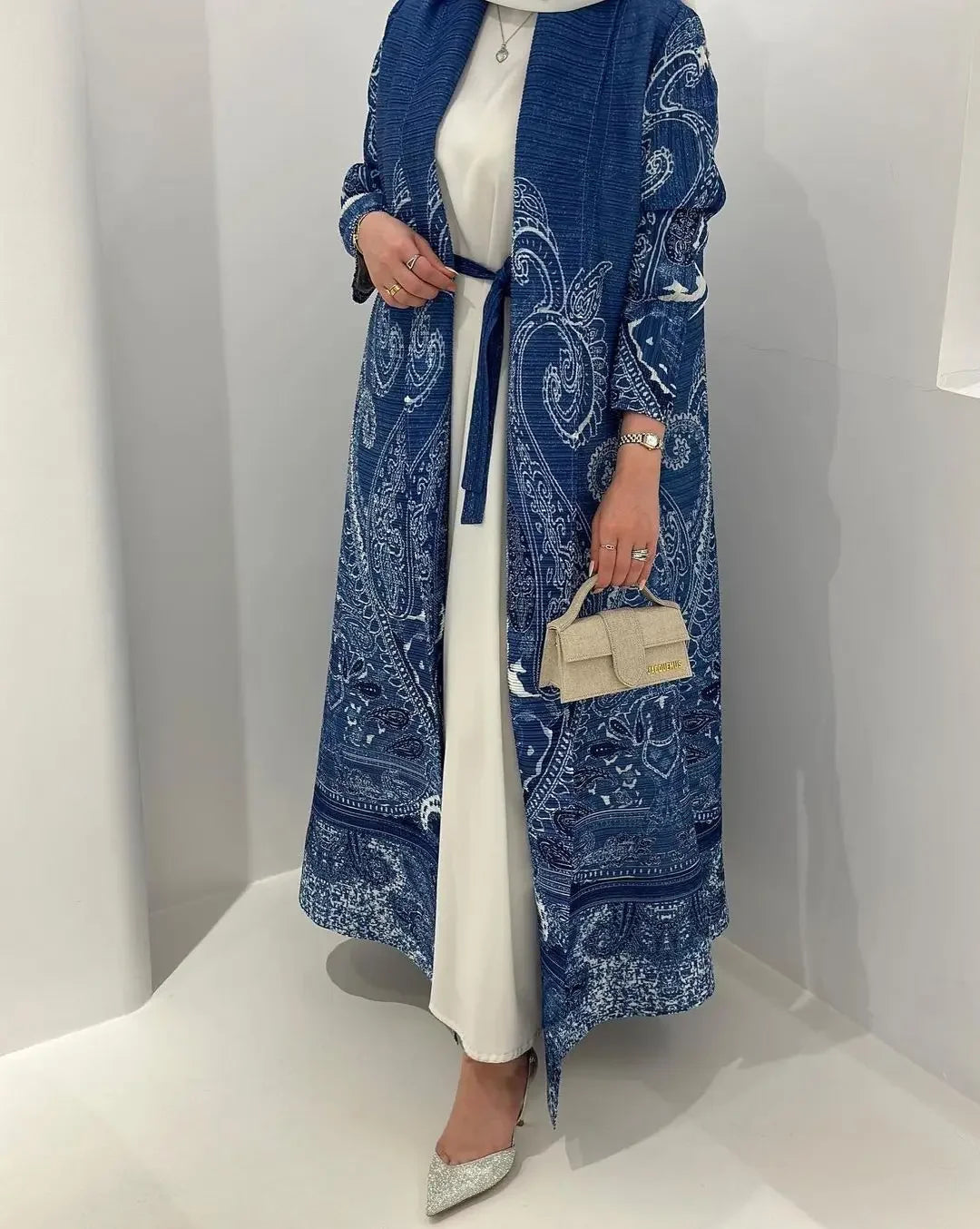 Une femme modélise une veste plissée bleu et blanc sur une robe blanche dans une galerie lumineuse.