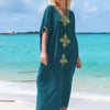 Eine Frau in einem türkisblauen Kaftan mit goldenen Stickereien steht am Strand bei klarem türkisfarbenem Wasser.