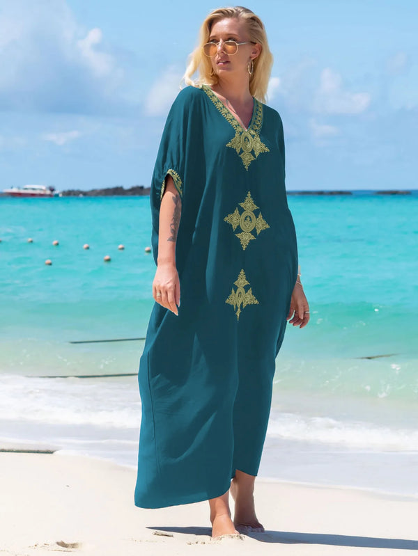 A woman wearing a teal kaftan dress with gold embroidery stands on a beach near clear turquoise water.