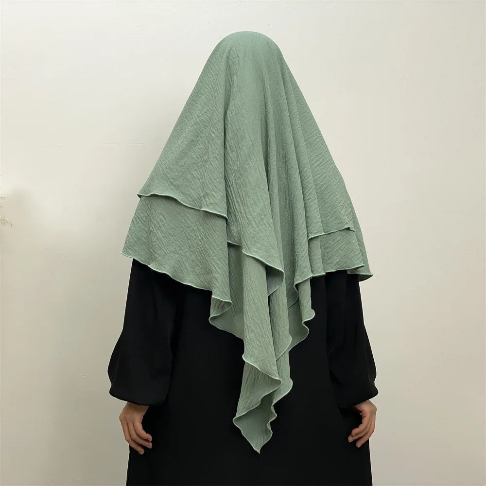 A woman wearing a light sage green two-layer everyday hijab scarf over a black top.