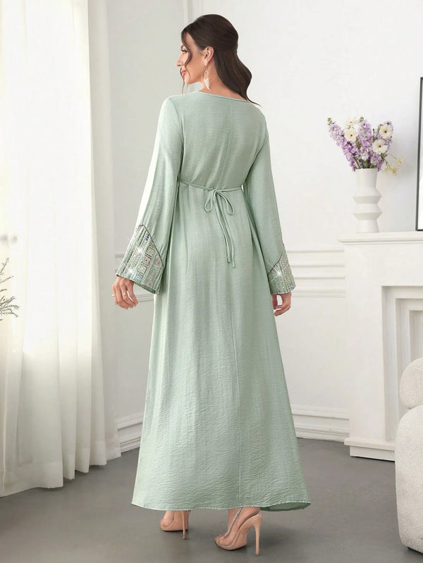 A woman models a light green abaya with sequin embroidery and a tie belt in a full-length shot.