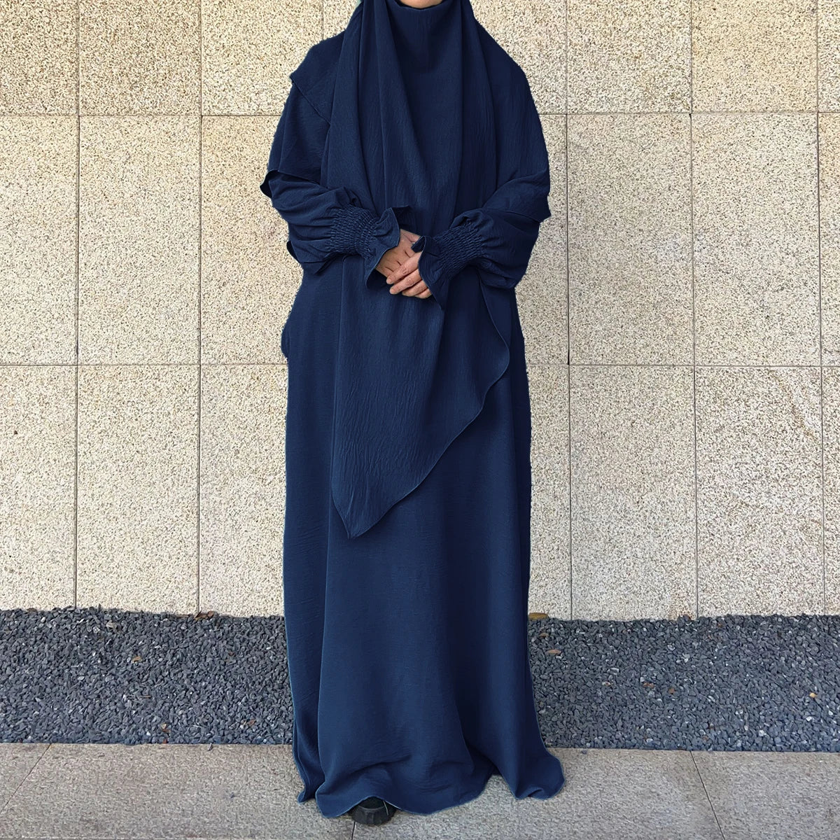 A dark blue elegant two-piece abaya and khimar set worn by a person against a light stone wall.
