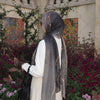 Eine Frau in grauem Hijab und weißer Kleidung steht inmitten üppiger lila und grüner Vegetation.