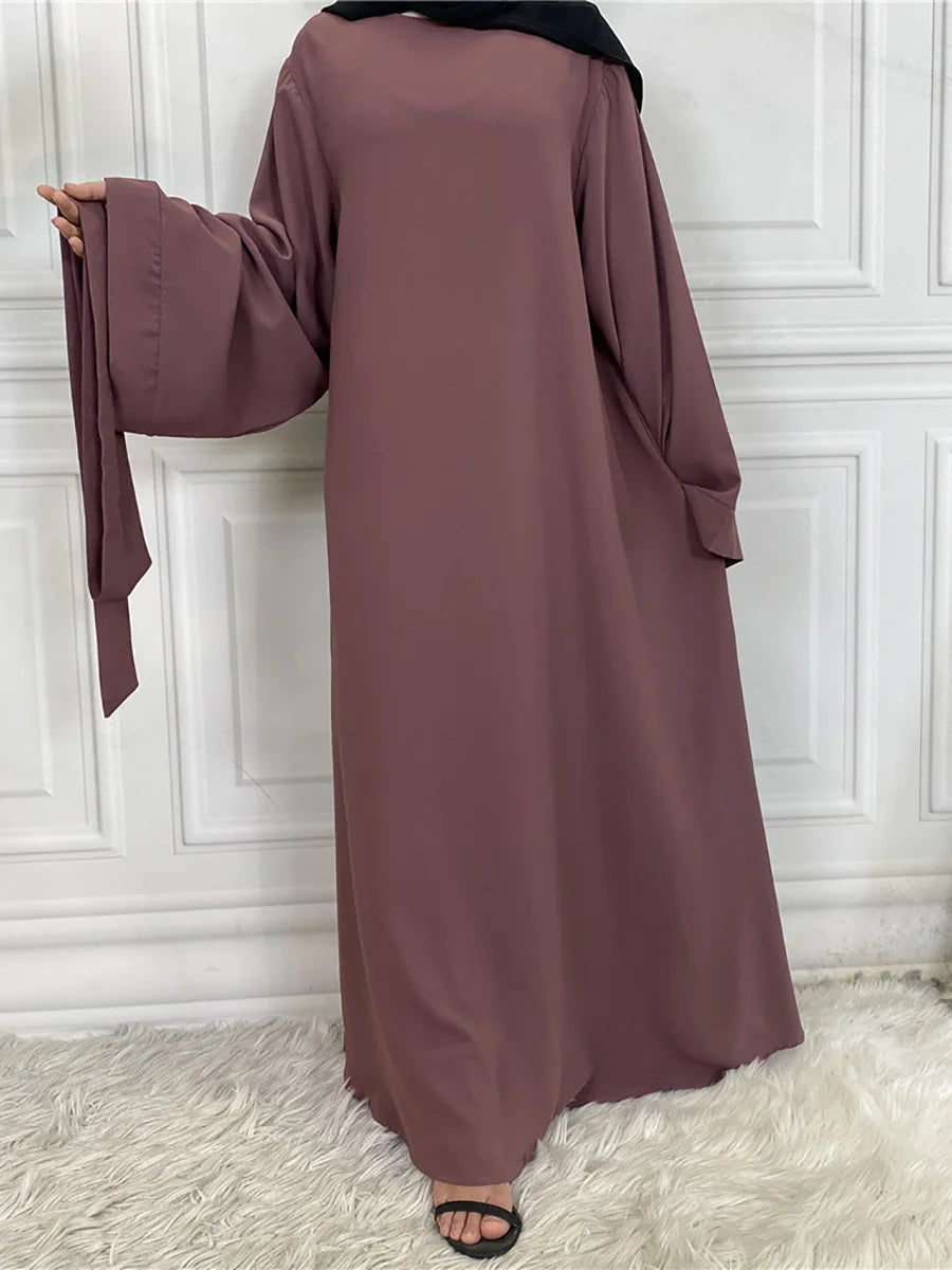 Maroon maxi abaya for women with long sleeves and a black hijab, displayed against a white paneled wall.
