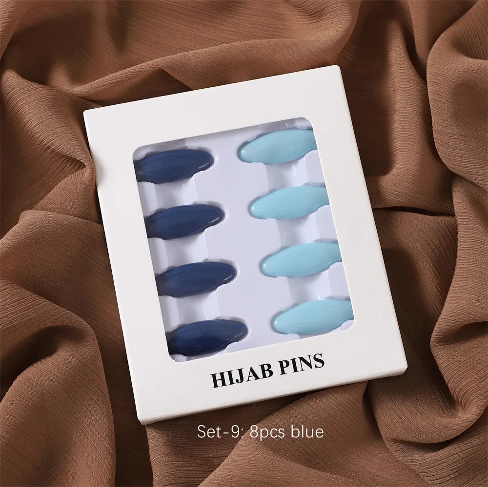 Ein Set von neun blauen Hijab-Pins in einer weißen quadratischen Box auf einem braunen Stoffhintergrund.