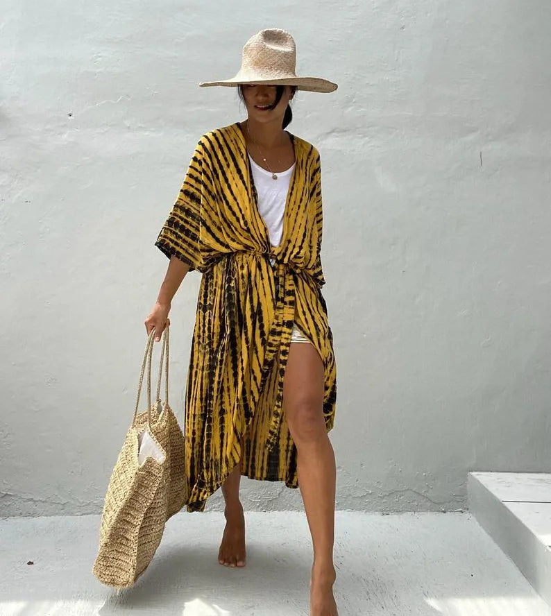 Eine Frau in einem gelb-schwarz gestreiften Sommer-Strandkimono-Kleid und einer Strohtasche vor einem hellgrauen Hintergrund