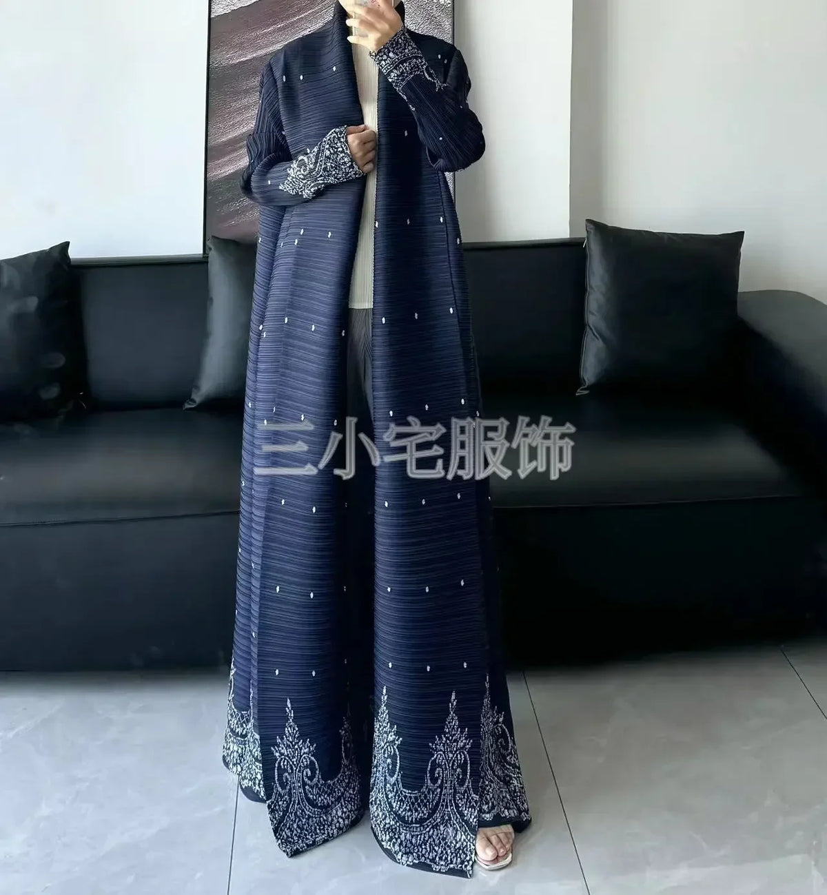 Une femme modélise un trench-coat à manches longues, plissé et à imprimé bleu foncé avec broderie argentée, dans un salon mod