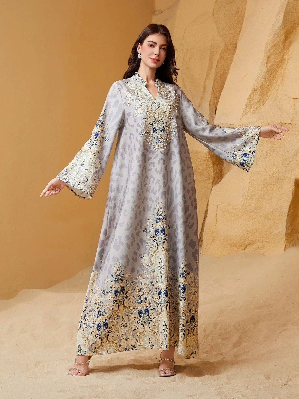 Una mujer modela una abaya larga y fluida de leopardo color marfil y azul con adornos de diamantes contra un fondo arenoso.