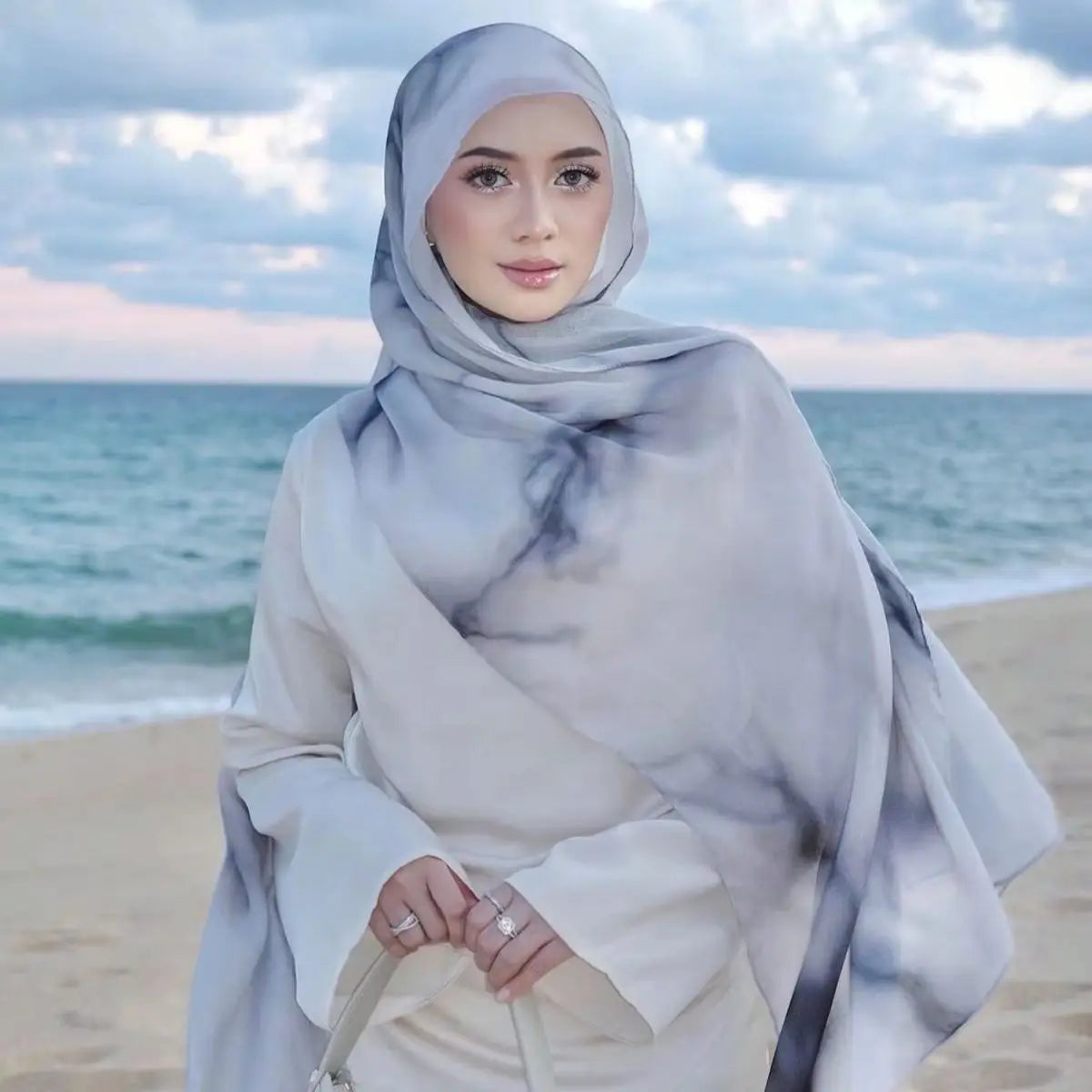 Eine Frau trägt einen eleganten bedruckten Baumwoll-Rayon-Hijab in hellgrauen und weißen Tönen am Strand.