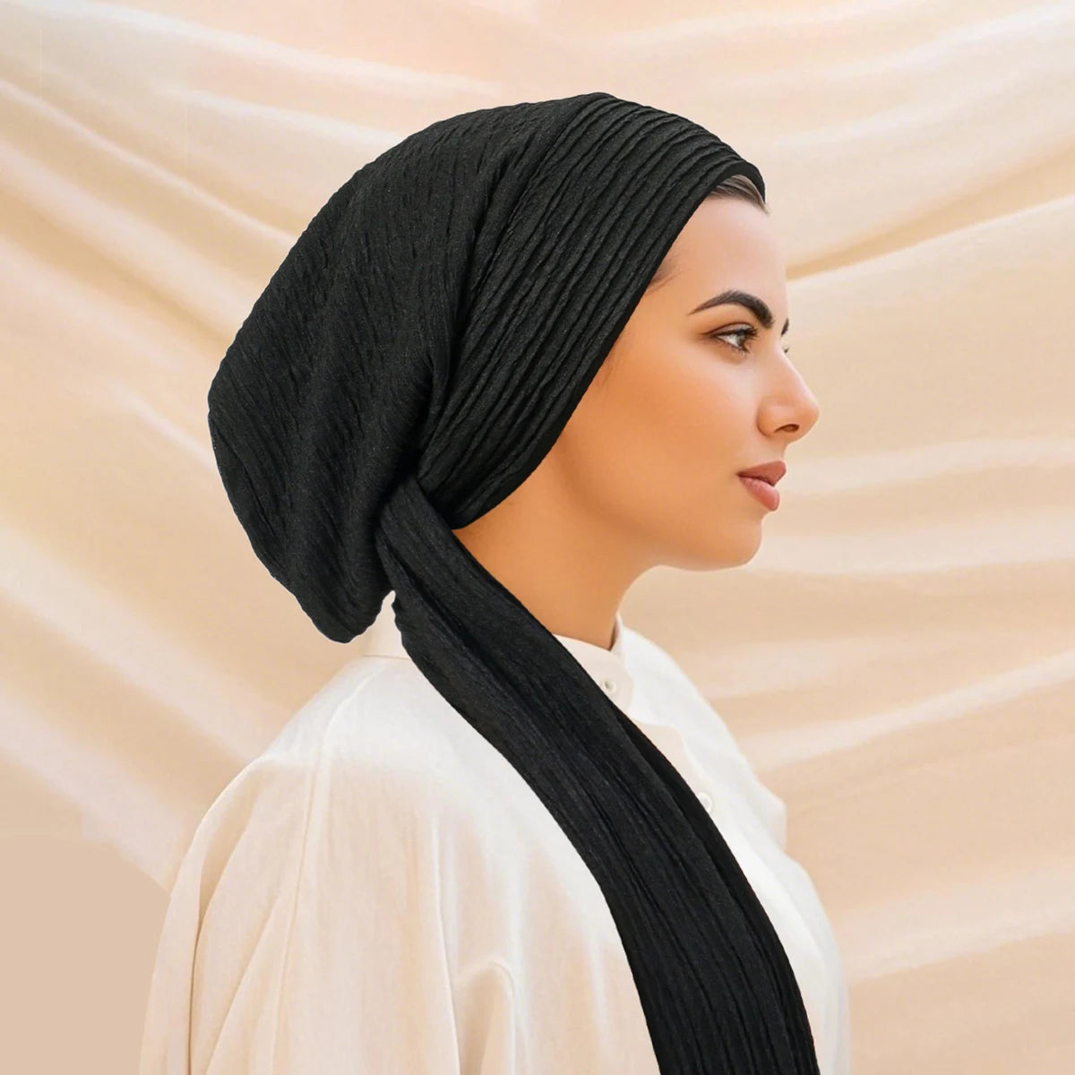 A woman wearing a solid black turban hijab cap and a white top, shown in profile against a light background.