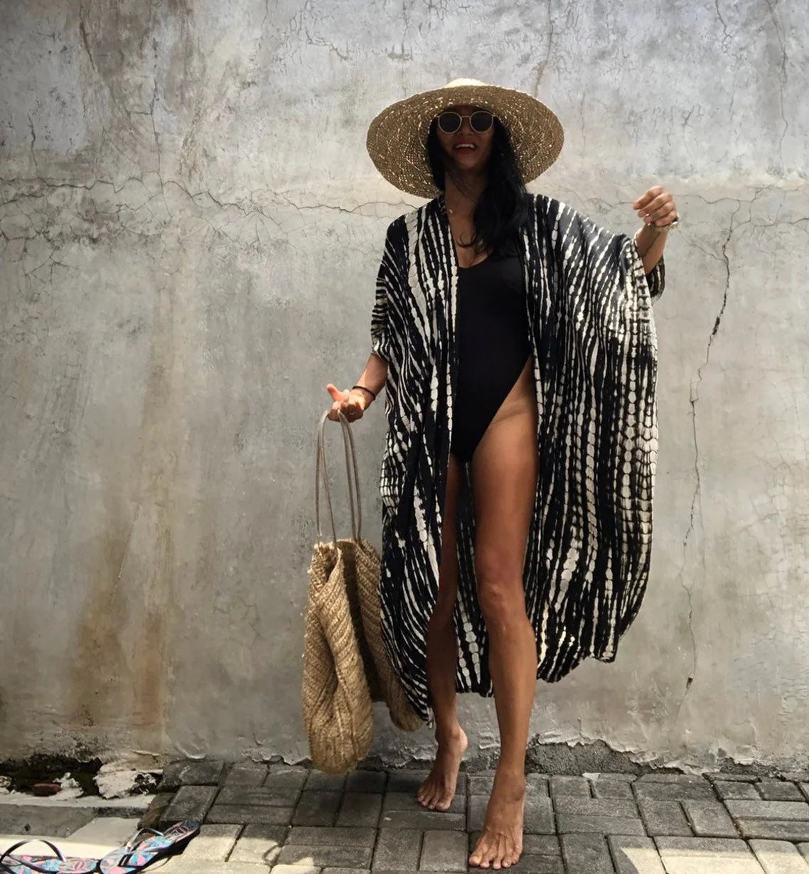 Eine Frau in einem schwarzen Badeanzug und einem gemusterten Kimono-Überkleid mit Strohhut vor einer Betonwand.