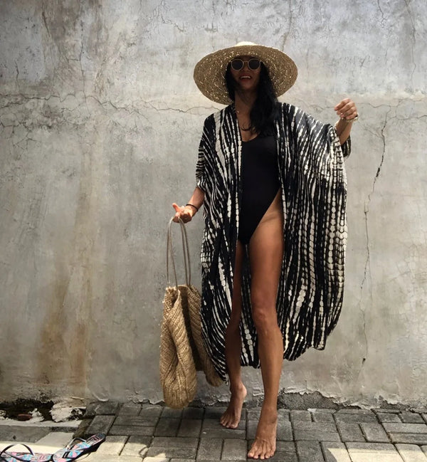 A woman wearing a black swimsuit and a patterned kimono cover-up dress with a straw hat standing against a concrete wall.
