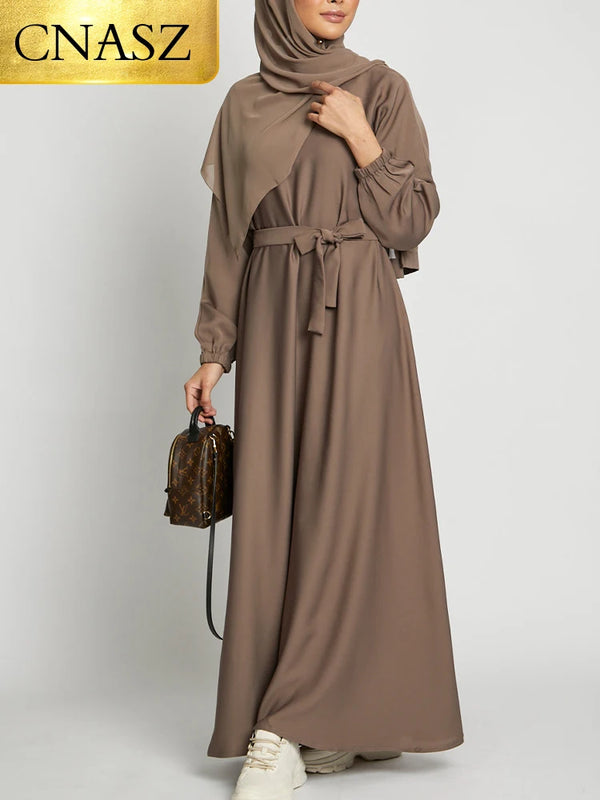 A woman wearing a long, flowing taupe Moroccan style kaftan abaya with long sleeves and a belted waist.