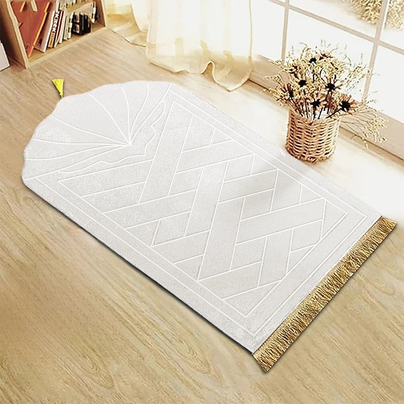 Alfombra de oración blanca grabada con patrón geométrico y borlas decorativas doradas sobre un suelo de madera claro.